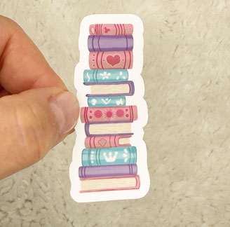 A hand holding a colorful sticker shaped like stacked books.