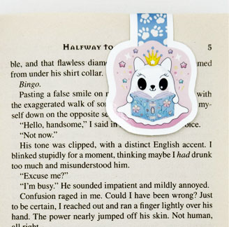 An illustrated magnetic bookmark featuring a cute white cat wearing a crown and reading a book, placed in a book.